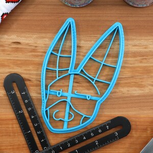 May include: A blue plastic cookie cutter shaped like a rabbit's head with a detailed design. The cutter is on a wooden surface.