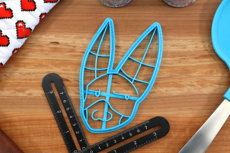 May include: A blue plastic cookie cutter shaped like a rabbit's head with a detailed design. The cutter is on a wooden surface.