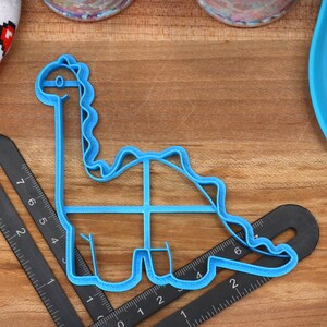 May include: Blue plastic dinosaur cookie cutter on a wooden surface with a ruler.