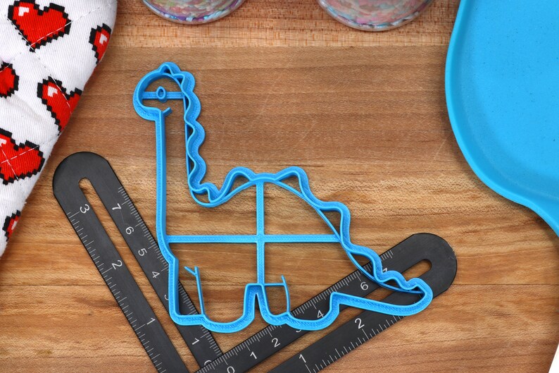 May include: Blue plastic dinosaur cookie cutter on a wooden surface with a ruler.
