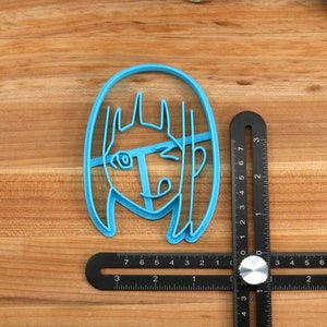 May include: A blue plastic cookie cutter shaped like a cartoon girl with long hair. The cutter is on a wooden surface with a black ruler.