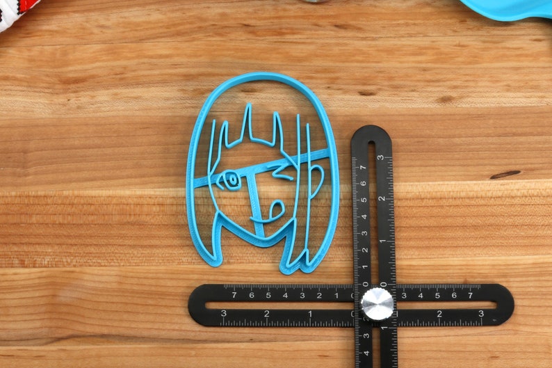 May include: A blue plastic cookie cutter shaped like a cartoon girl with long hair. The cutter is on a wooden surface with a black ruler.