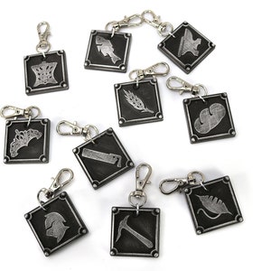 May include: Ten silver keychains with different symbols on black square tags. The symbols include a crown, a fish, an anvil, a wheat stalk, a leaf, a saw, a helmet, a pickaxe, and two more leaves.