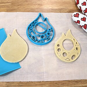 May include: Two cookie cutters in the shape of a flame, one blue and one beige, with a beige cookie dough cutout on a white parchment paper surface.
