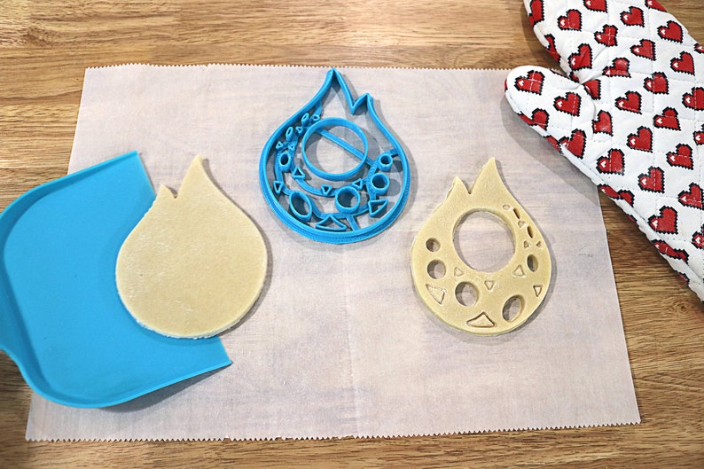 May include: Two cookie cutters in the shape of a flame, one blue and one beige, with a beige cookie dough cutout on a white parchment paper surface.