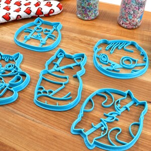 May include: Five blue plastic cookie cutters shaped like cats. The cutters are on a wooden surface.