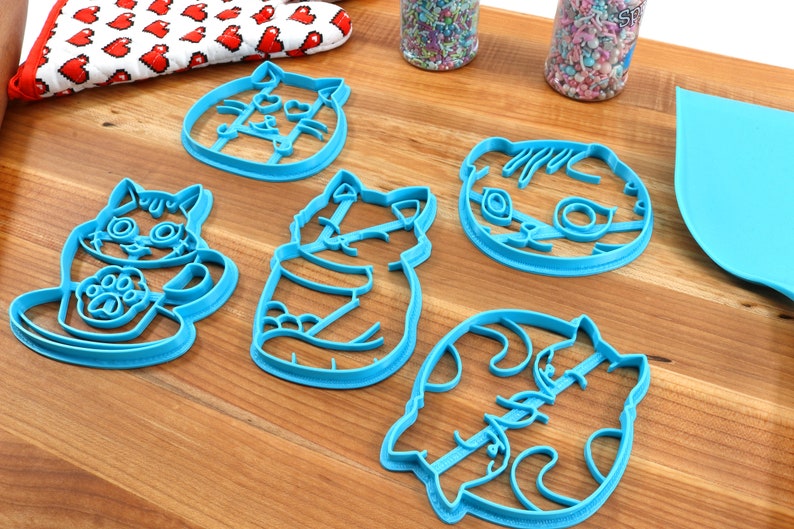 May include: Five blue plastic cookie cutters shaped like cats. The cutters are on a wooden surface.