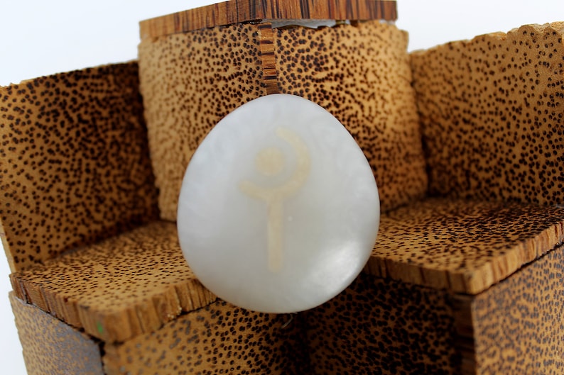 May include: A white egg-shaped stone with a white symbol etched into the surface. The stone is resting on a wooden box with a brown and black speckled pattern.