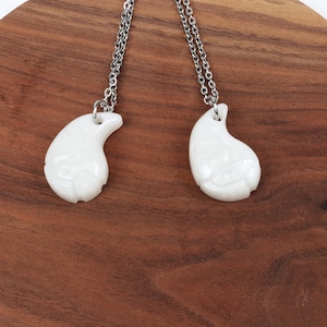 May include: Two white teardrop-shaped pendants on silver chains. The pendants have a carved design and are connected together.