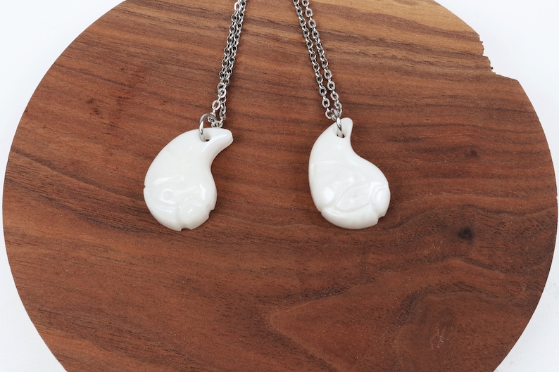 May include: Two white teardrop-shaped pendants on silver chains. The pendants have a carved design and are connected together.