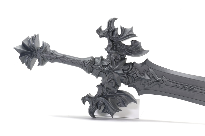 May include: A detailed, ornate, silver-colored sword with a decorative hilt and a sharp blade. The sword is displayed on a white stand.