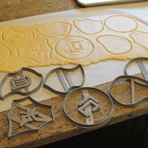 May include: A sheet of cookie dough with various shapes cut out using cookie cutters. The cookie cutters are shown on a counter top next to the dough.