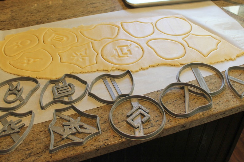May include: A sheet of cookie dough with various shapes cut out using cookie cutters. The cookie cutters are shown on a counter top next to the dough.