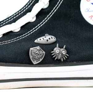 May include: Black Converse high-top trainers with white laces and a white star logo. Three silver metal pins are attached to the shoe: a shield with a bird design, a mask with spikes, and a teardrop-shaped object with holes.