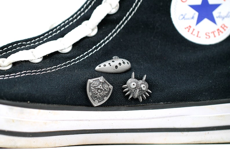 May include: Black Converse high-top trainers with white laces and a white star logo. Three silver metal pins are attached to the shoe: a shield with a bird design, a mask with spikes, and a teardrop-shaped object with holes.