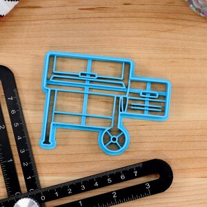 May include: A blue plastic cookie cutter shaped like a grill with a wheel on one side. The cookie cutter is on a wooden surface.