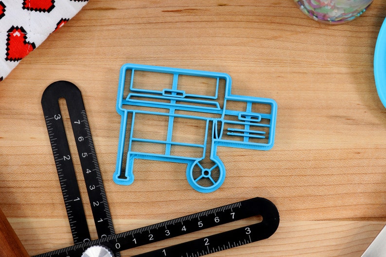 May include: A blue plastic cookie cutter shaped like a grill with a wheel on one side. The cookie cutter is on a wooden surface.