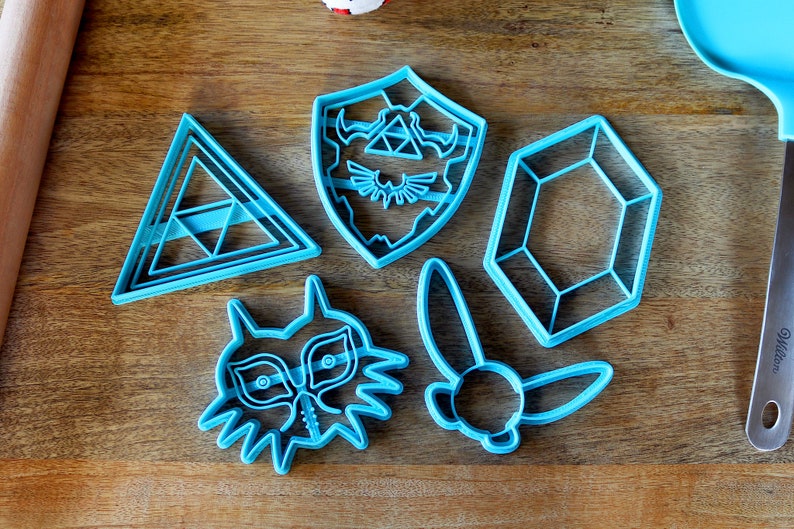 Puede incluir: Cinco cortadores de galletas de pl&aacute;stico azul con forma de escudo con un s&iacute;mbolo Triforce, un diamante, un tri&aacute;ngulo, una cabeza de lobo estilizada y un p&aacute;jaro estilizado con alas.