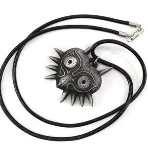 May include: A black and silver pendant necklace featuring a stylized heart-shaped mask with sharp points and two large eyes. The necklace is made of a black cord.