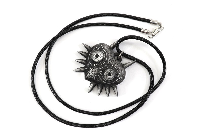 May include: A black and silver pendant necklace featuring a stylized heart-shaped mask with sharp points and two large eyes. The necklace is made of a black cord.