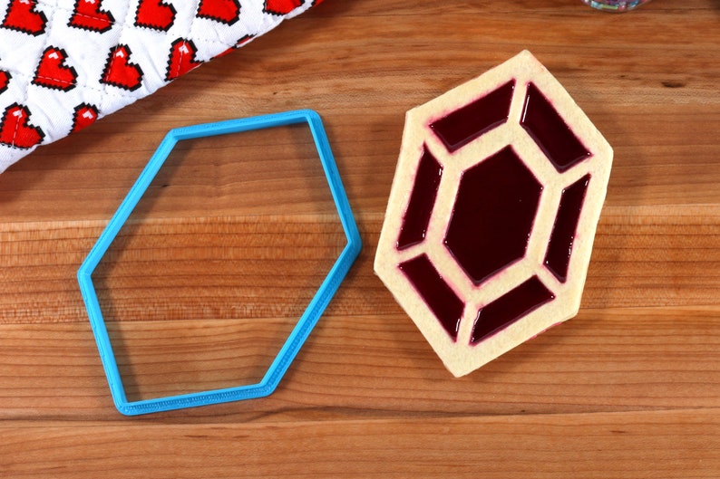 Op de afbeelding: Een blauwe plastic koekjesvormer in de vorm van een zeshoek, en een koekje in de vorm van een zeshoek met een rode gelei vulling.