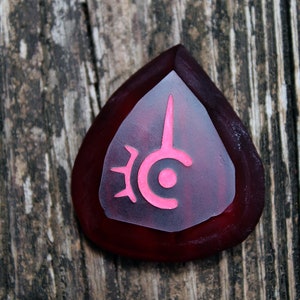 May include: A red guitar pick with a pink symbol on it. The symbol is a stylized circle with a spike coming out of the top.