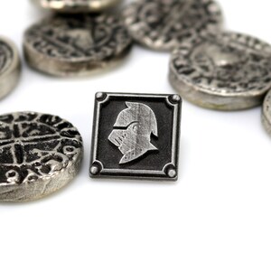 May include: A silver metal pin with a black square frame and a silver knight's helmet design. The pin is surrounded by several silver metal coins with intricate designs.