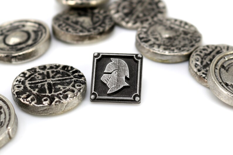 May include: A silver metal pin with a black square frame and a silver knight's helmet design. The pin is surrounded by several silver metal coins with intricate designs.
