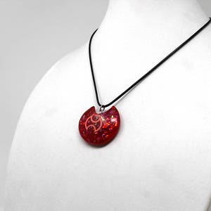 May include: A red and pink glitter resin pendant necklace with a black cord. The pendant is shaped like a crescent moon with a stylized symbol in the center.