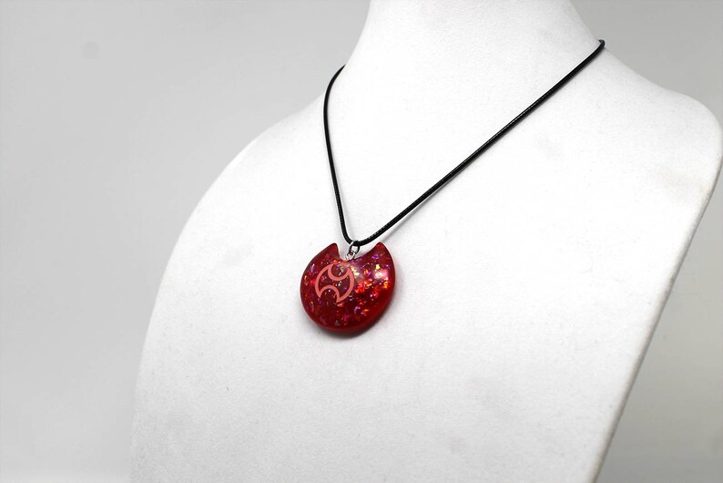 May include: A red and pink glitter resin pendant necklace with a black cord. The pendant is shaped like a crescent moon with a stylized symbol in the center.