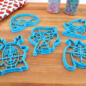 Peut inclure: Ensemble de d&eacute;coupoirs &agrave; biscuits en plastique bleu en forme de personnages de dessins anim&eacute;s, dont un renne, un chat et un personnage avec des lunettes. Un gant de cuisine rouge et blanc &agrave; motifs de c&oelig;urs et deux pots de vermicelles sont &eacute;galement visibles.