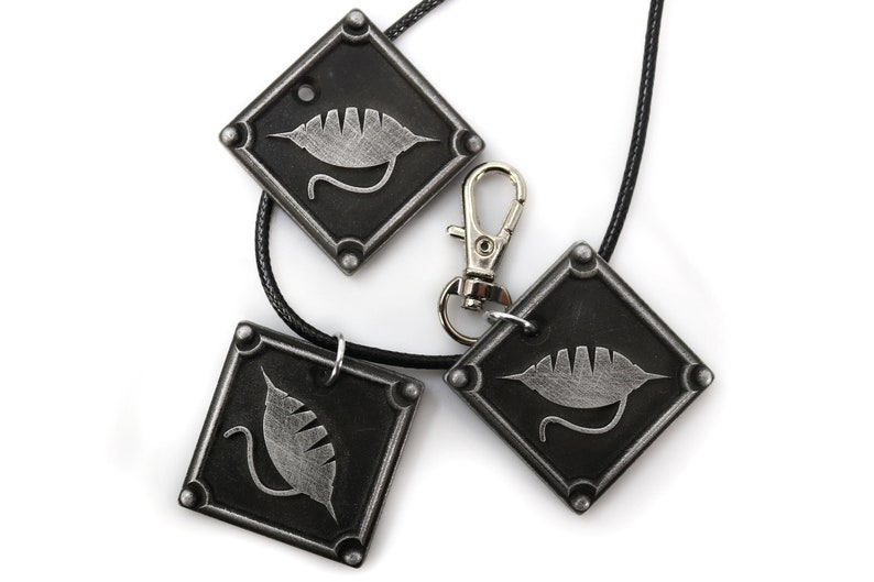 Puede incluir: Tres colgantes de metal negro con un dise&ntilde;o de hoja plateada. Cada colgante tiene forma de diamante y est&aacute; unido a un cord&oacute;n negro. Los colgantes est&aacute;n sobre un fondo blanco.