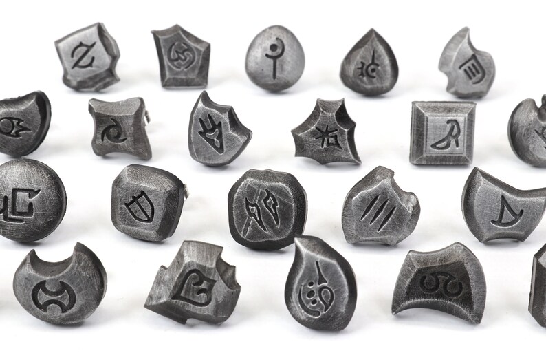 Puede incluir: Una colecci&oacute;n de veintiuna piezas de juego de metal gris desgastado con s&iacute;mbolos grabados. Las piezas tienen formas variadas, incluyendo cuadrados, c&iacute;rculos y formas irregulares. Cada pieza presenta un dise&ntilde;o grabado &uacute;nico.