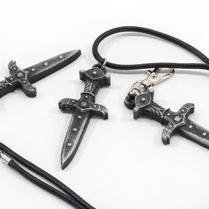 May include: Three silver-colored dagger pendants with intricate details. Two of the pendants are attached to black cords, while the third is lying on a white surface.