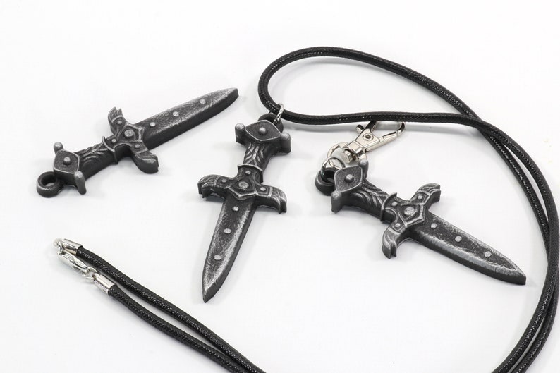 May include: Three silver-colored dagger pendants with intricate details. Two of the pendants are attached to black cords, while the third is lying on a white surface.