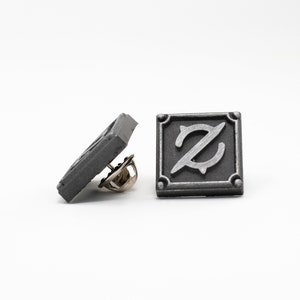 Op de afbeelding: Een zilverkleurige metalen pin met een vierkant ontwerp met een gestileerde letter Z in het midden. De pin heeft een veiligheidssluiting aan de achterkant.