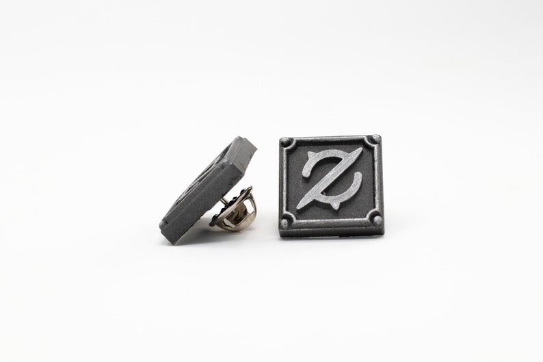 Op de afbeelding: Een zilverkleurige metalen pin met een vierkant ontwerp met een gestileerde letter Z in het midden. De pin heeft een veiligheidssluiting aan de achterkant.