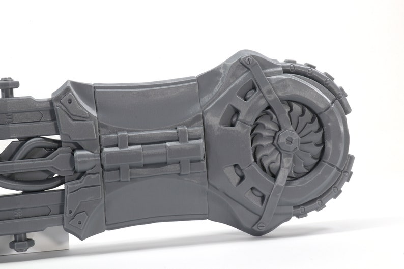 May include: A gray 3D printed model of a futuristic weapon with a large, detailed gear on the right side.