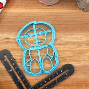 May include: A blue plastic cookie cutter shaped like a cartoon character with glasses. The character has a round head, a wide smile, and is wearing a shirt with a button.