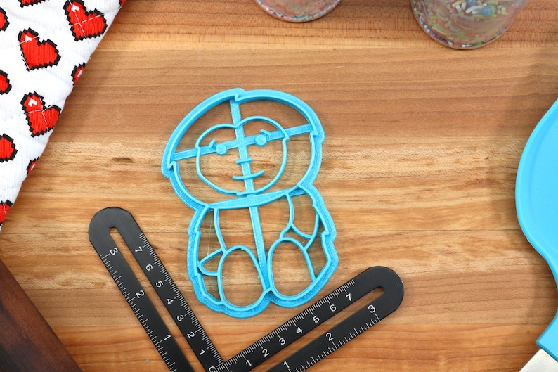 May include: A blue plastic cookie cutter shaped like a cartoon character with glasses. The character has a round head, a wide smile, and is wearing a shirt with a button.
