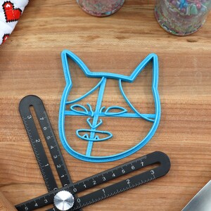 May include: A blue plastic cookie cutter shaped like a cat's face. The cutter has a simple design with a line for the nose and eyes, and a curved line for the mouth.
