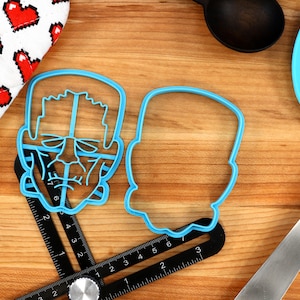 May include: Two blue plastic cookie cutters shaped like Frankenstein's face. The cutter on the left has a detailed face with eyes, nose, and mouth. The cutter on the right is a simple outline of a head.