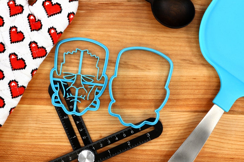 May include: Two blue plastic cookie cutters shaped like Frankenstein's face. The cutter on the left has a detailed face with eyes, nose, and mouth. The cutter on the right is a simple outline of a head.