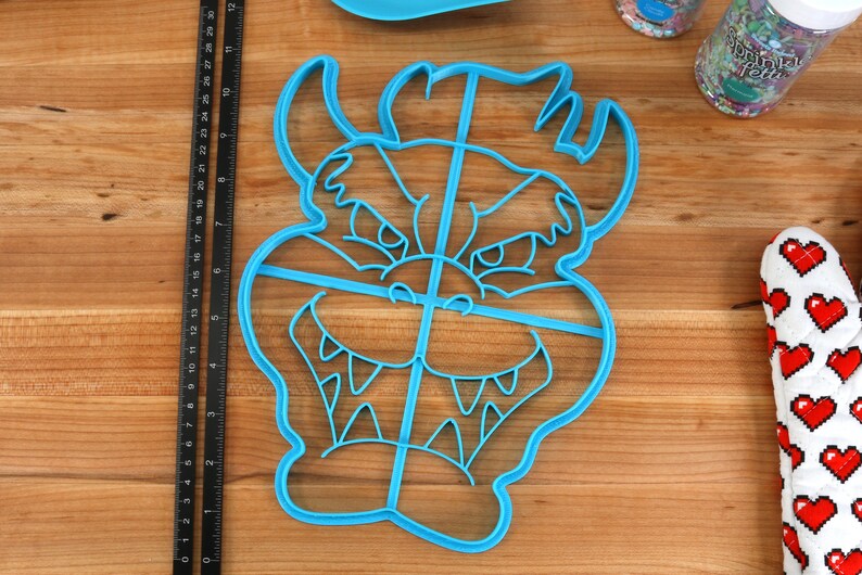 Op de afbeelding: Blauwe plastic koekjesvormer in de vorm van een cartoon monster gezicht met een wijd open mond en scherpe tanden. De vormer is verdeeld in vier secties door lijnen.