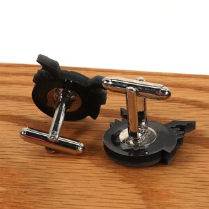 May include: A pair of black cufflinks with a silver metal clasp. The cufflinks feature a stylized design of a winged creature.