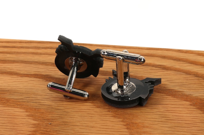 May include: A pair of black cufflinks with a silver metal clasp. The cufflinks feature a stylized design of a winged creature.