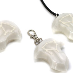 Op de afbeelding: Drie witte hangers met een sterrenbeeldontwerp, een aan een zwarte koordsnoer, een aan een zilveren sleutelhanger en een los. Het sterrenbeeld is Weegschaal.