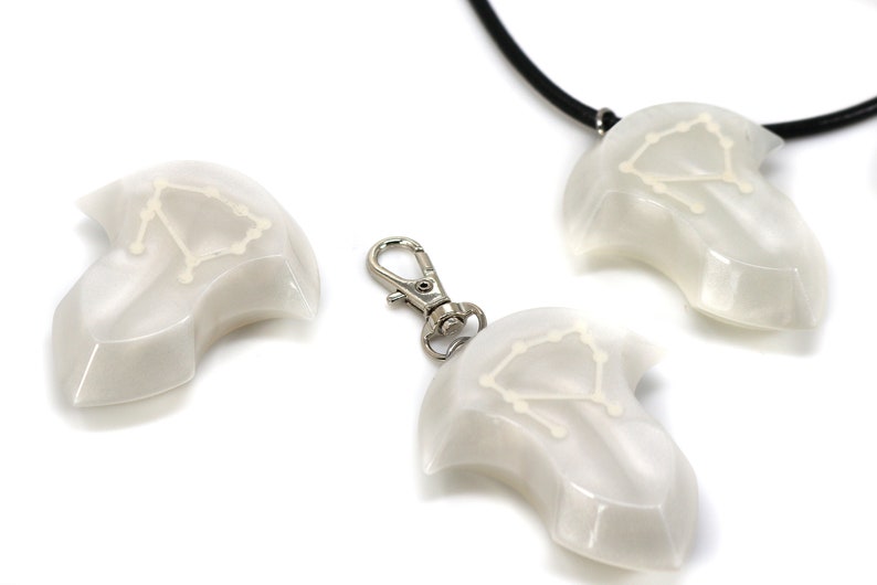 Op de afbeelding: Drie witte hangers met een sterrenbeeldontwerp, een aan een zwarte koordsnoer, een aan een zilveren sleutelhanger en een los. Het sterrenbeeld is Weegschaal.