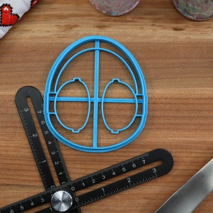May include: A blue plastic cookie cutter shaped like an egg with two smaller circles inside. The cutter is on a wooden surface with a ruler and a spatula.