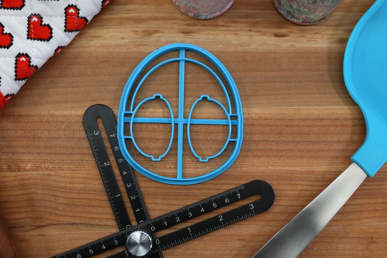 May include: A blue plastic cookie cutter shaped like an egg with two smaller circles inside. The cutter is on a wooden surface with a ruler and a spatula.